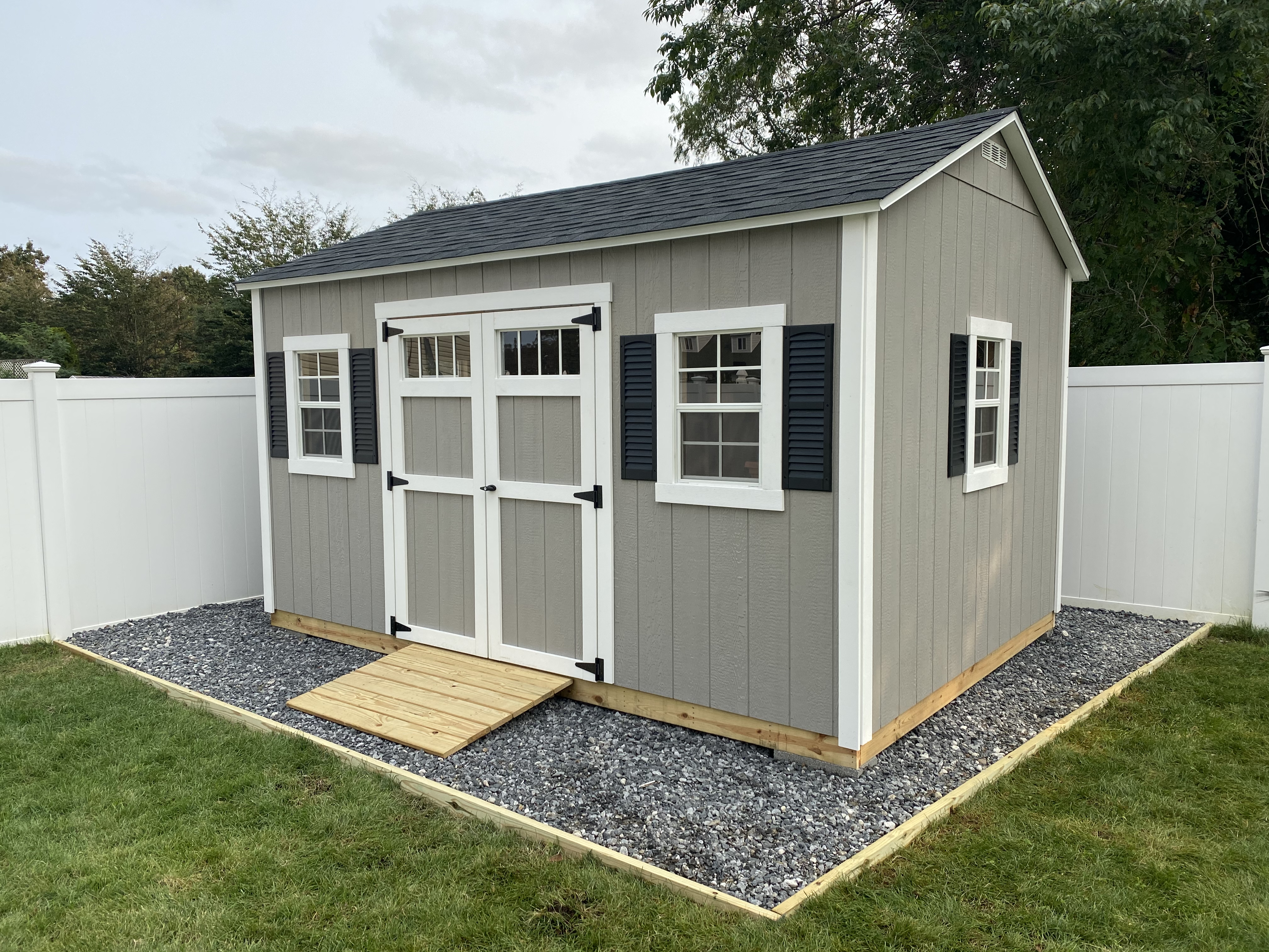 Beige gable-roof storage shed with white trim, double barn doors, two front windows with black shutters, and a wooden entry ramp.