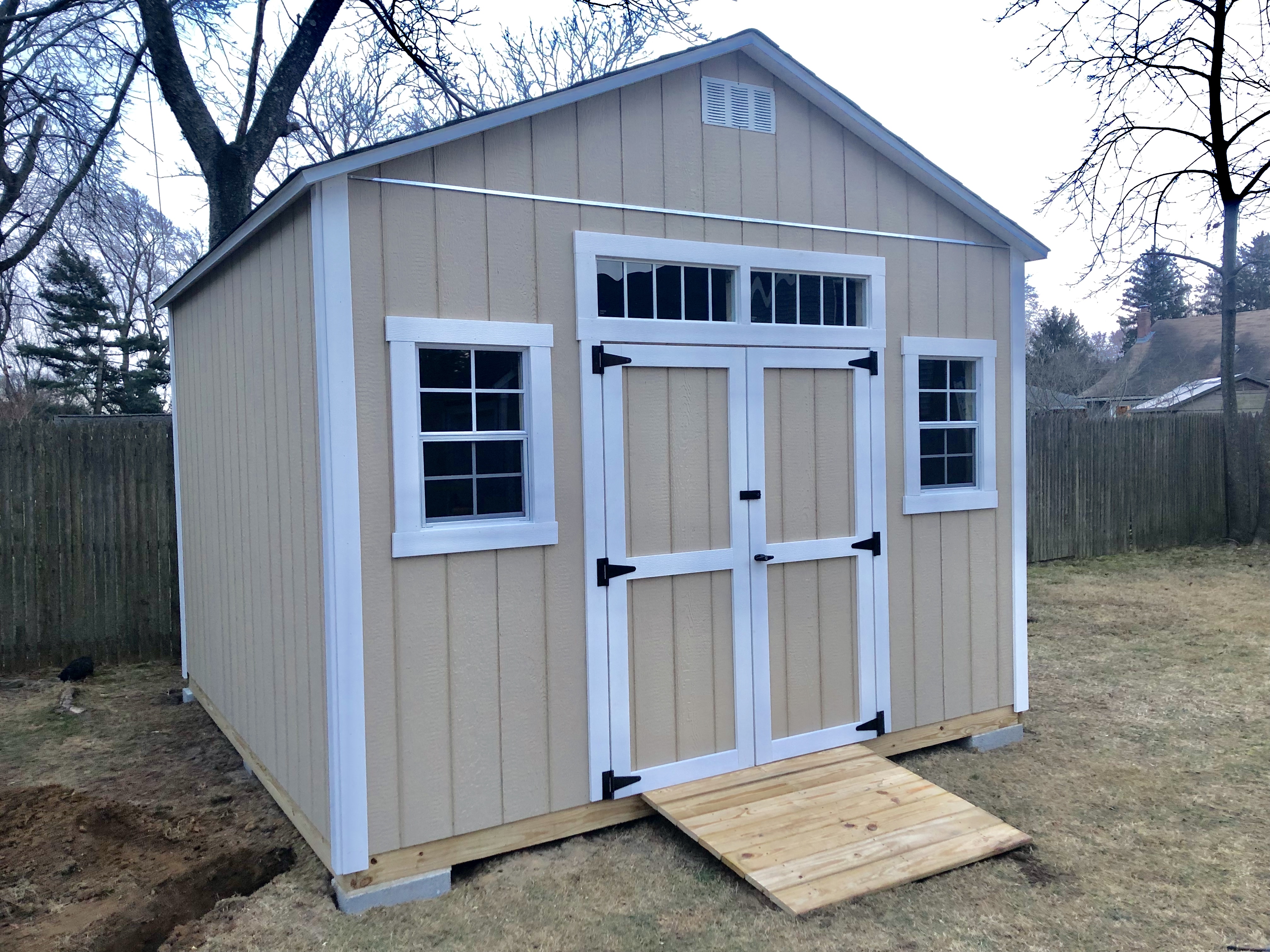 Tall tan gable-roof storage shed with white trim, double barn doors, and a decorative transom window above the doors.