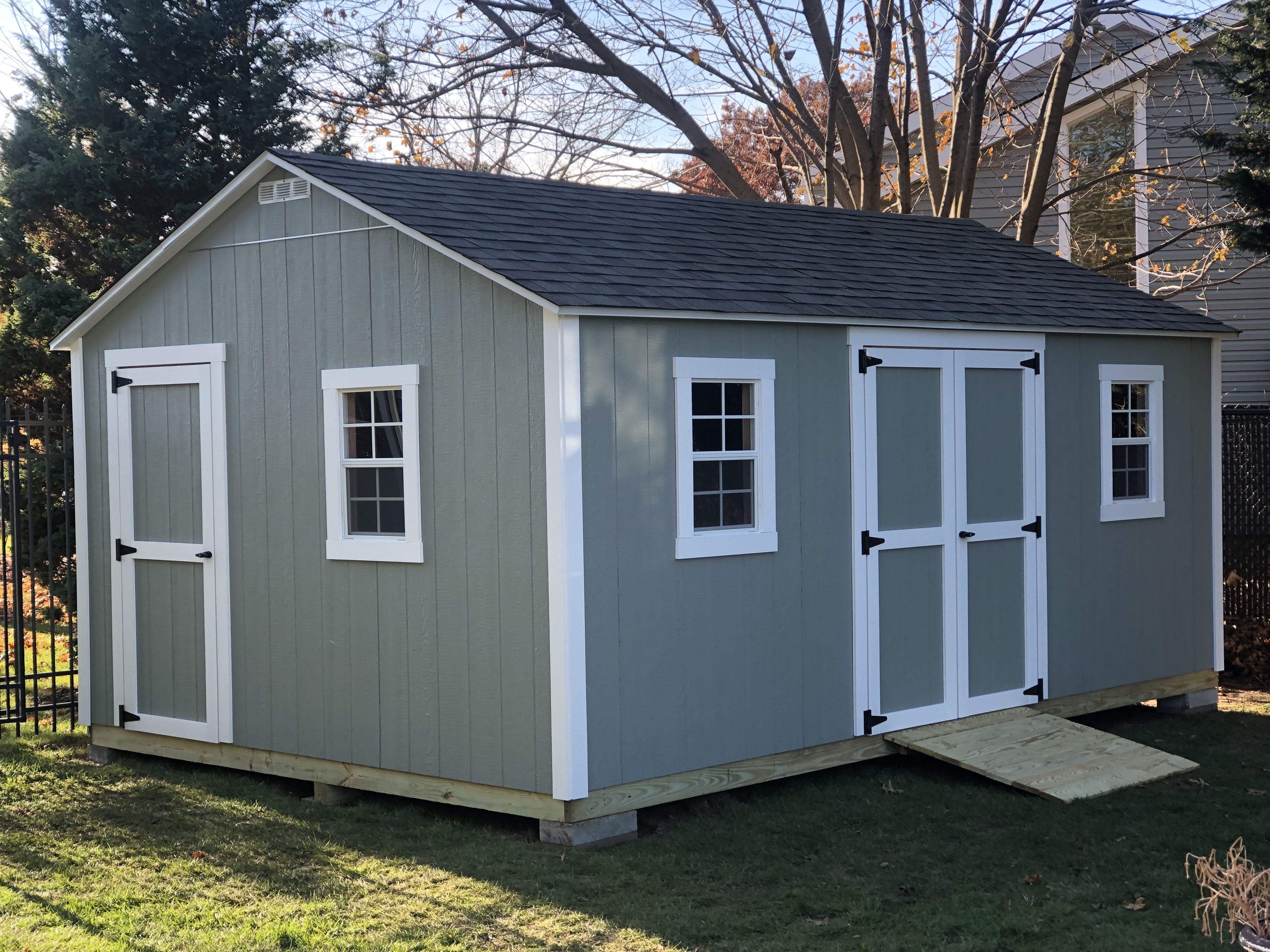 Sage green gable-roof storage shed with white trim, double doors, and two front windows.