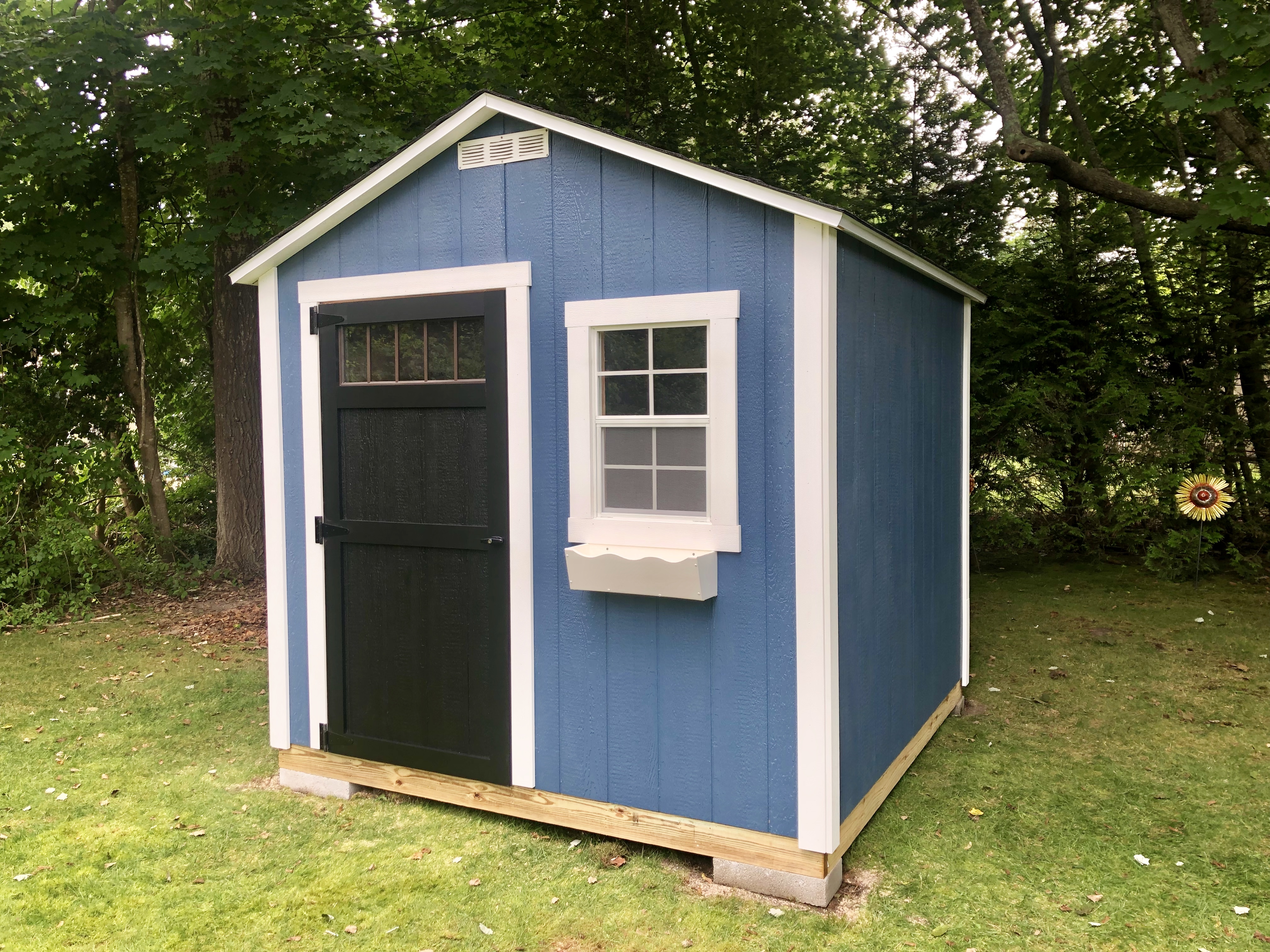 Blue garden shed with white trim, a black single door, and a front window with a decorative flower box.