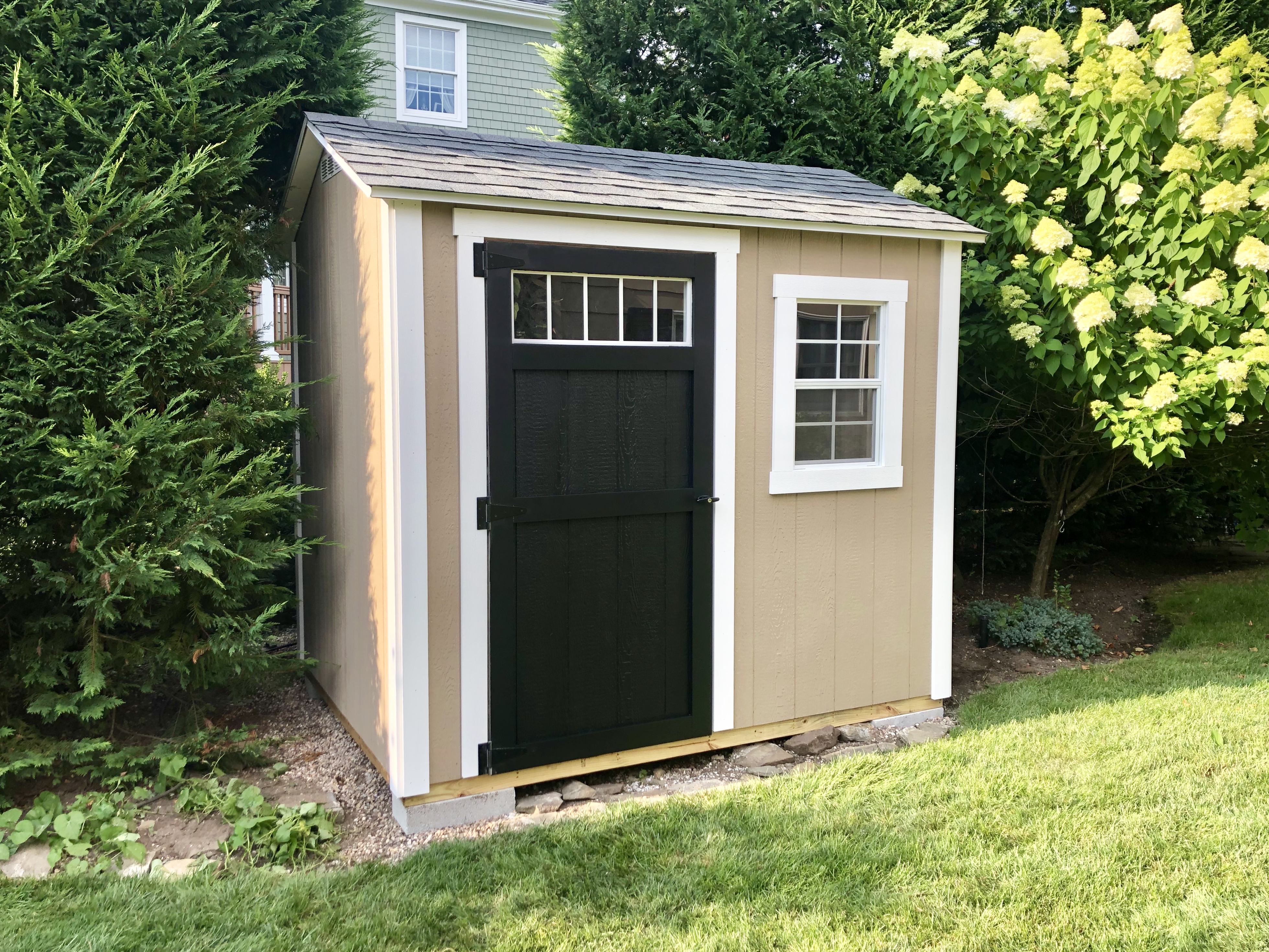 Small tan garden shed with white trim, a dark green single door, and a front window.