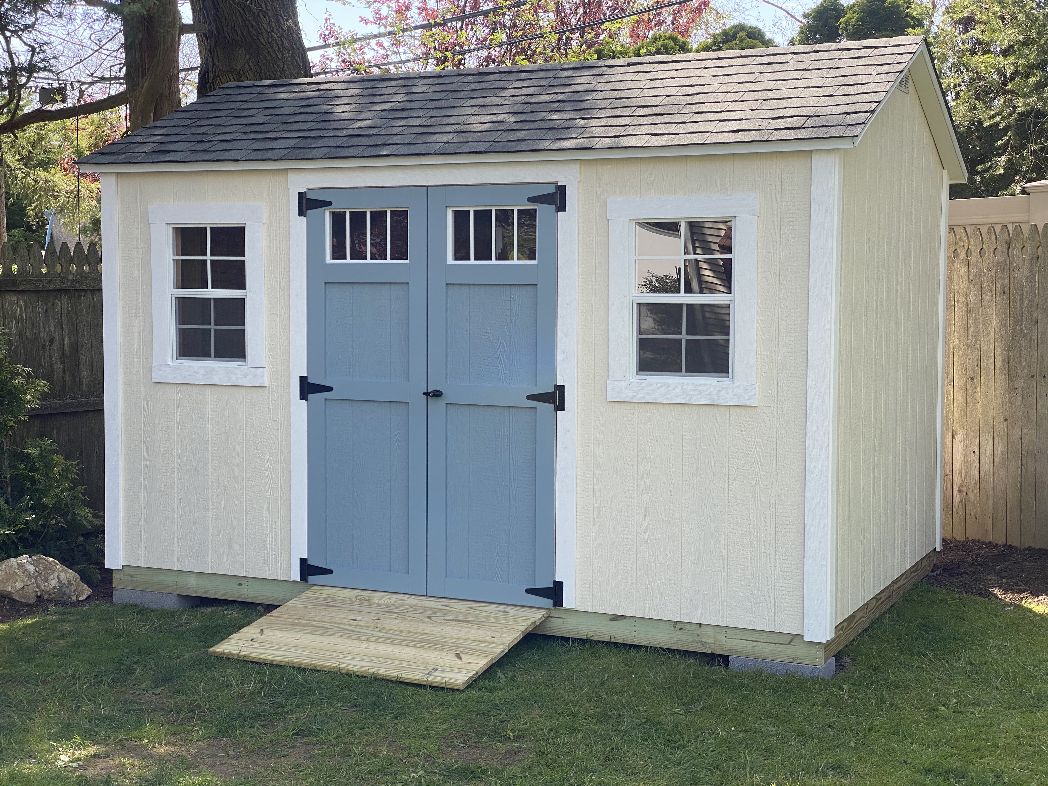 Cream colored gable-roof storage shed with white trim and blue carriage-style double doors with windows.